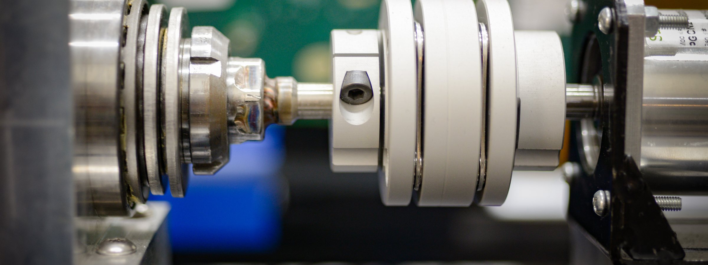 Close-up of a bench rig: a white flexible shaft coupling with a hex set screw joins a polished steel shaft to a stack of precision bearings, with a green circuit board and a blue fixture out of focus behind.