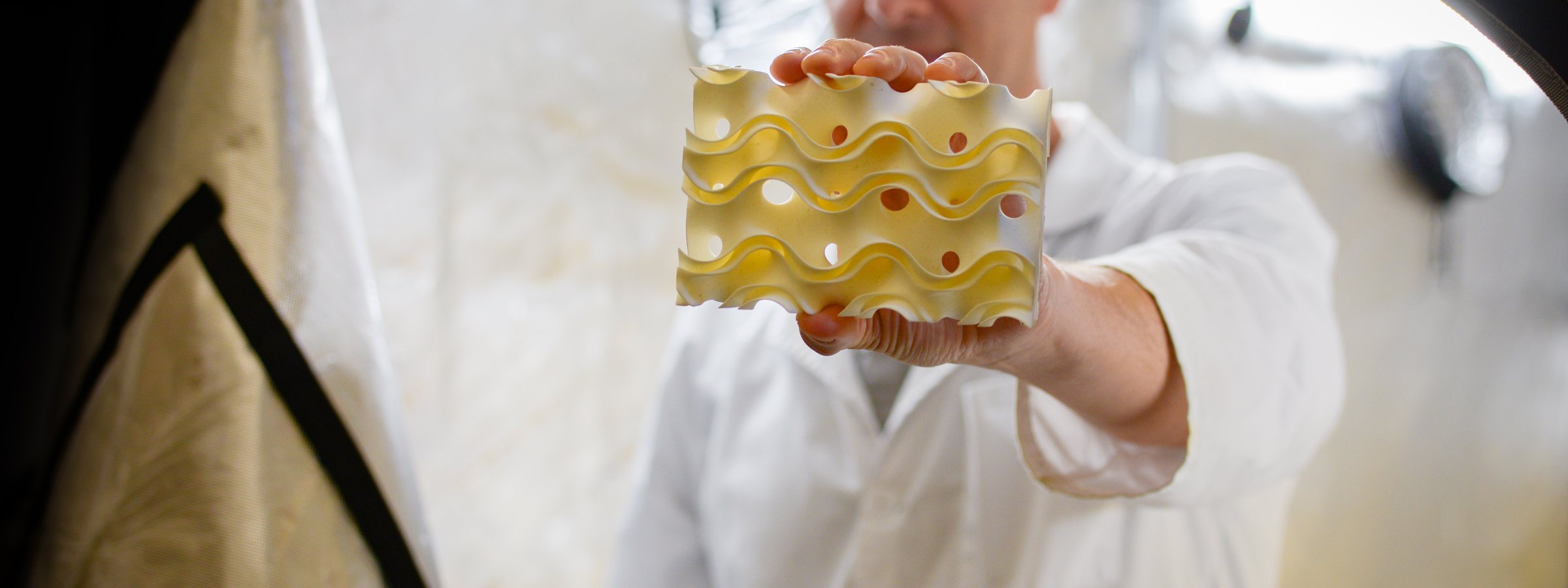Someone in a white lab coat holds a pale yellow 3D-printed block out towards the camera: stacked wavy fins with round holes through them, in a bright workshop.
