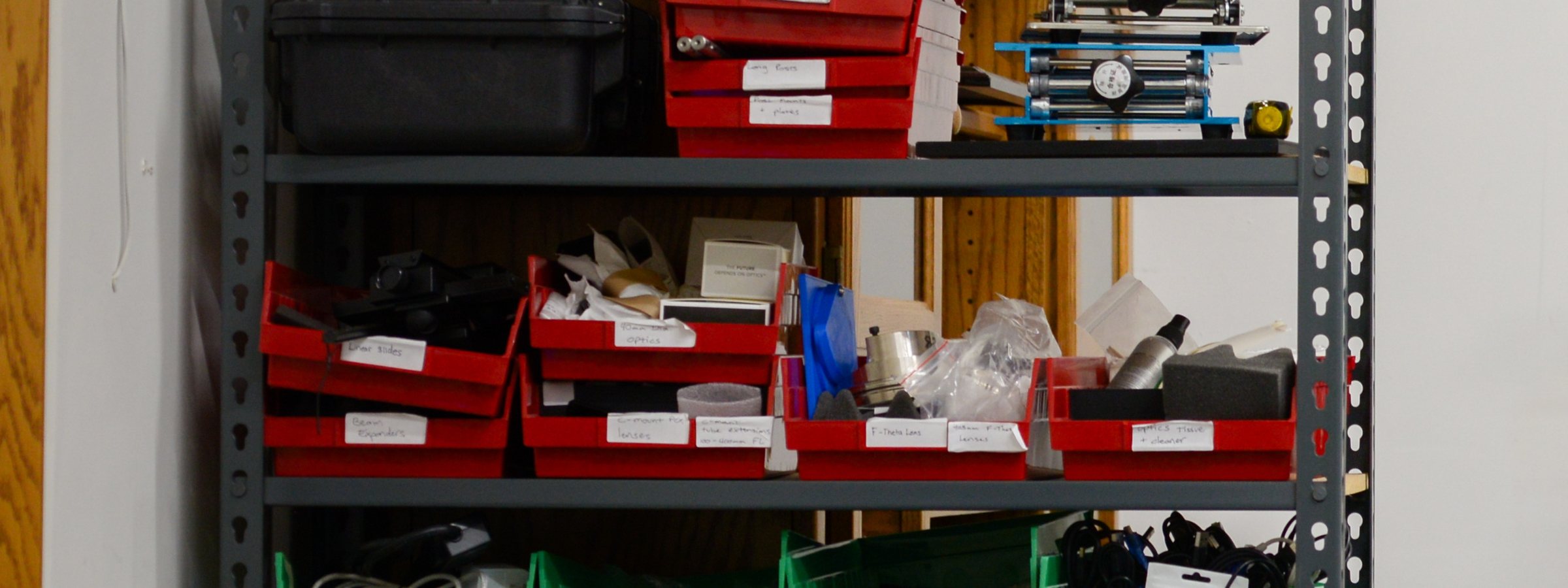 Grey steel shelving in the shop, stacked with red and green parts bins hand-labelled &lsquo;linear slides&rsquo;, &lsquo;beam expanders&rsquo;, &lsquo;F-theta lens&rsquo; and &lsquo;optics tissue&rsquo;, alongside cased equipment and coiled cables.