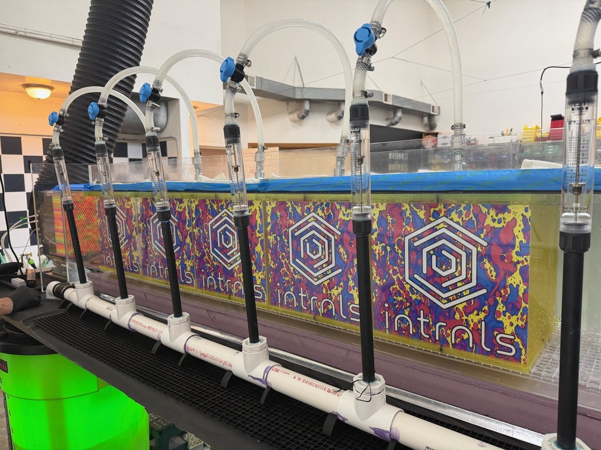 A long clear-walled water tank on a bench, printed media stacked inside it, with six flow meters on a PVC manifold feeding tubes down through the lid; the tank panel is printed with the intrnls wordmark.