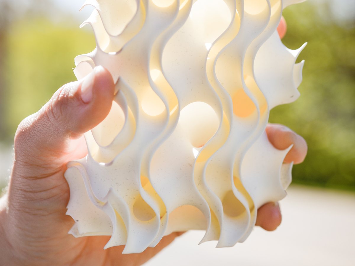 A hand holds up a white 3D-printed block of stacked wavy fins; sunlight behind it glows through the curved channels and the round openings cut into them.